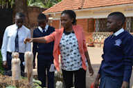 A teacher explaining a concept to three students in school uniforms outdoors, with a focus on a hands-on activity involving plastic bottles and a garden setup. The background shows a brick building and greenery.